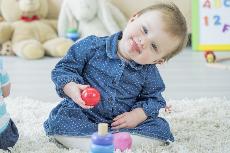 A baby girl is sitting on the carpet and making a funny face while playing with toys